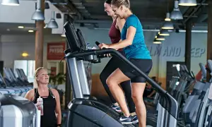 Två kvinnor tränar på trappmaskiner. En tredje kvinna med vattenflaska tittar på. Gym maskiner namn är inte synliga, men det är en modern gymmiljö.