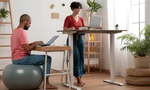 Man sitter på en träningsboll och jobbar vid laptop, medan en kvinna står upp vid ett höj- och sänkbart skrivbord. Ett modernt sätt att stå upp på jobbet.