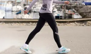 Kvinna i träningskläder går längs en kaj. Hon har på sig svarta tights och en lila tröja med texten "Running for Coke". Skorna är vita med blå detaljer. Hon tar många steg per km.
