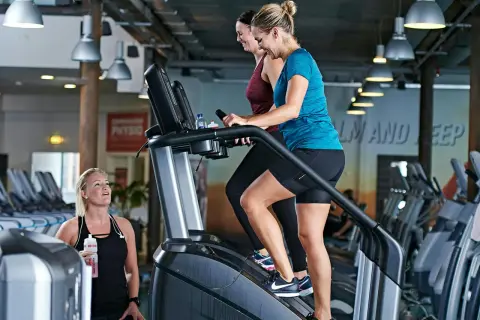 Två kvinnor tränar på trappmaskiner. En tredje kvinna med vattenflaska tittar på. Gym maskiner namn är inte synliga, men det är en modern gymmiljö.