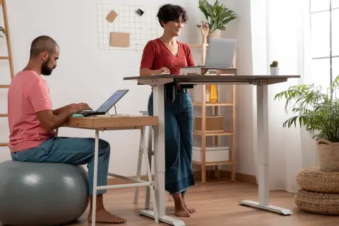 Man sitter på en träningsboll och jobbar vid laptop, medan en kvinna står upp vid ett höj- och sänkbart skrivbord. Ett modernt sätt att stå upp på jobbet.