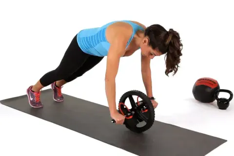 Kvinna utför ab wheel rollout på en träningsmatta. En kettlebell och en medicinboll syns i bakgrunden.
