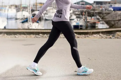 Kvinna i träningskläder går längs en kaj. Hon har på sig svarta tights och en lila tröja med texten "Running for Coke". Skorna är vita med blå detaljer. Hon tar många steg per km.