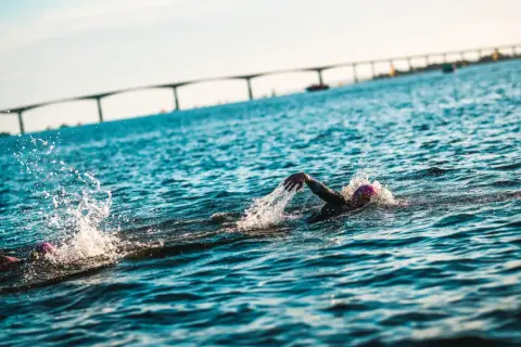 Simmare kämpar sig fram i vattnet under en ironman simning. En bro syns i bakgrunden.