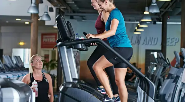 Två kvinnor tränar på trappmaskiner. En tredje kvinna med vattenflaska tittar på. Gym maskiner namn är inte synliga, men det är en modern gymmiljö.