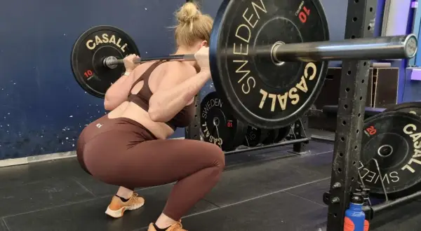En kvinna gör squats svenska med en skivstång på axlarna i ett gym.