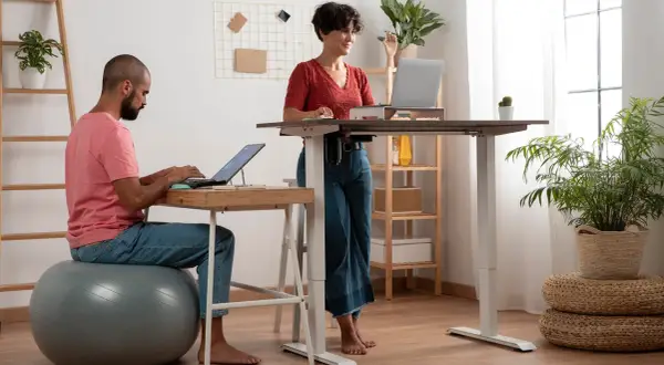Man sitter på en träningsboll och jobbar vid laptop, medan en kvinna står upp vid ett höj- och sänkbart skrivbord. Ett modernt sätt att stå upp på jobbet.