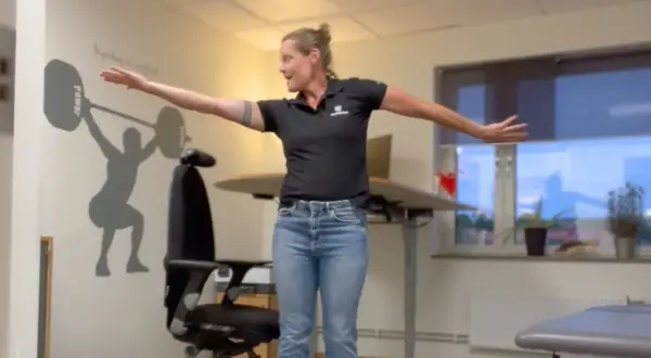 Kvinna utför en rörelse för att stretcha axel. Hon står framför en väggmålning av en tyngdlyftare.