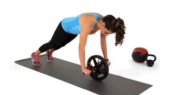 Kvinna utför ab wheel rollout på en träningsmatta. En kettlebell och en medicinboll syns i bakgrunden.