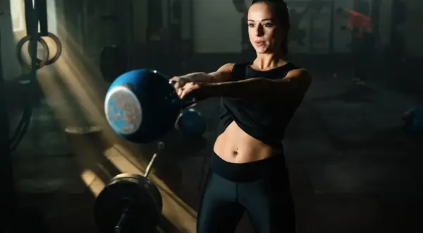 Kvinna utför marklyft med kettlebell i ett gym. Ljuset faller in från sidan och belyser hennes fokuserade ansikte och den blå kettlebellen.