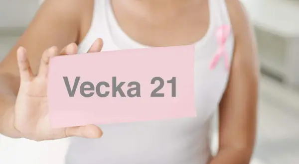 En person håller upp ett rosa kort med texten "Vecka 21". En rosa band pryder personens vita linne.