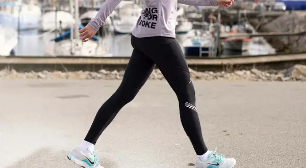 Kvinna i träningskläder går längs en kaj. Hon har på sig svarta tights och en lila tröja med texten "Running for Coke". Skorna är vita med blå detaljer. Hon tar många steg per km.
