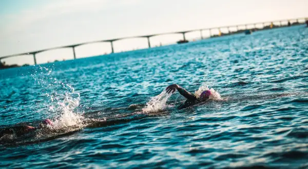 Simmare kämpar sig fram i vattnet under en ironman simning. En bro syns i bakgrunden.
