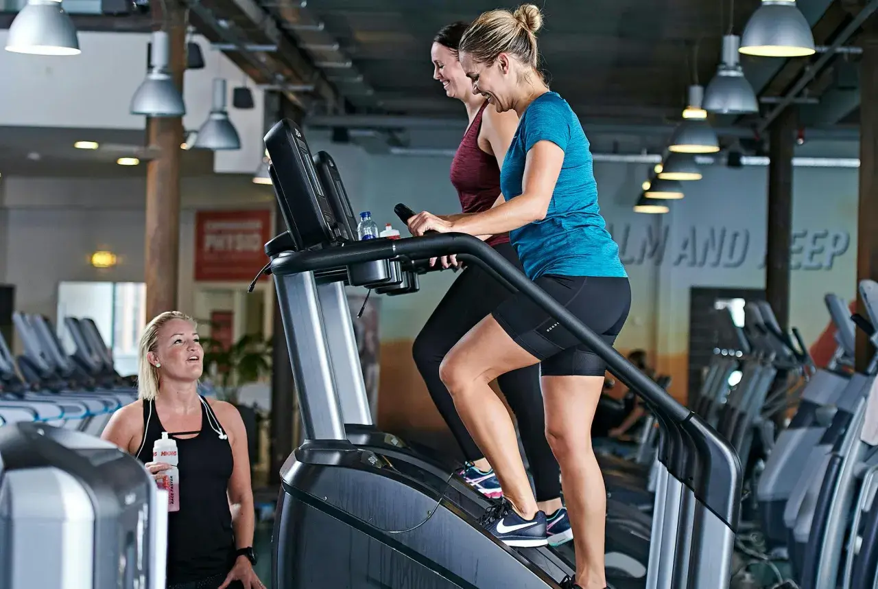 Två kvinnor tränar på trappmaskiner. En tredje kvinna med vattenflaska tittar på. Gym maskiner namn är inte synliga, men det är en modern gymmiljö.