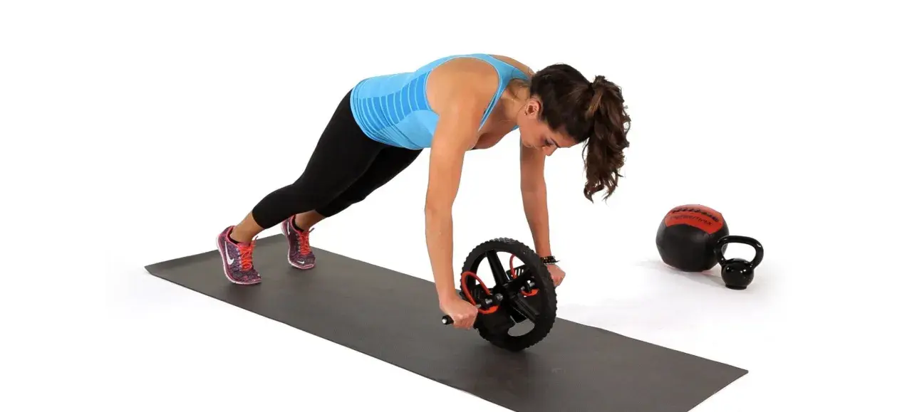 Kvinna utför ab wheel rollout på en träningsmatta. En kettlebell och en medicinboll syns i bakgrunden.