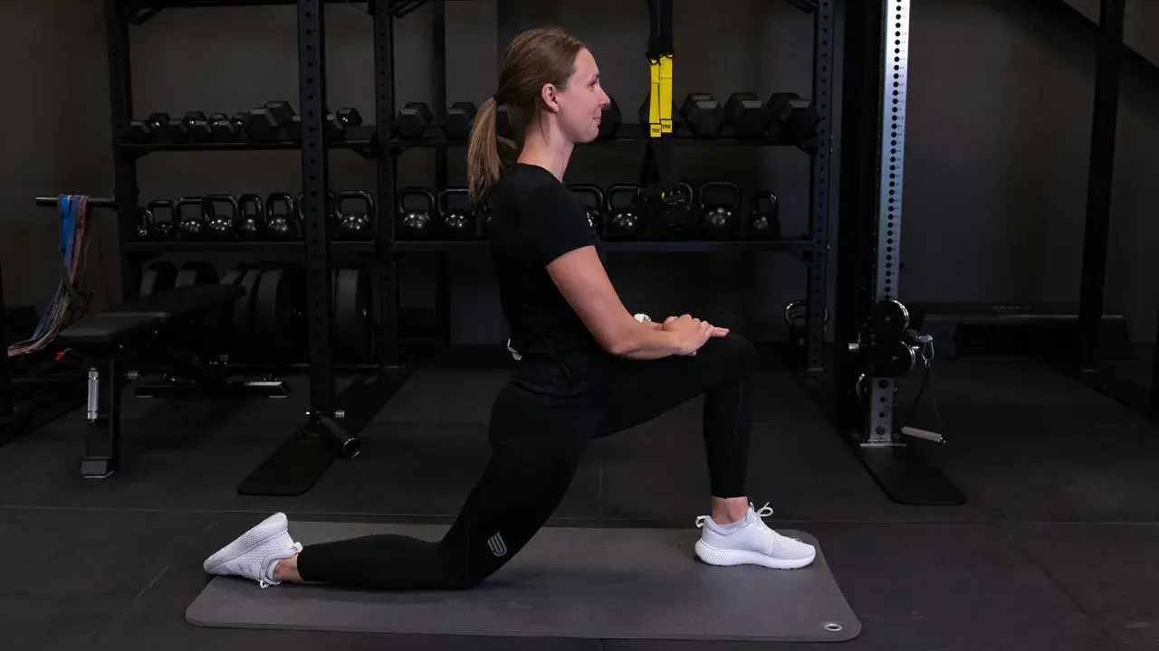 Kvinna utför grodan stretch på en träningsmatta i ett gym. Hantlar och vikter syns i bakgrunden.