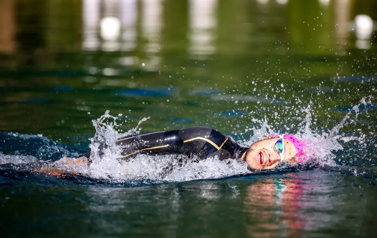 En t&auml;vlingssimmare i en v&aring;tdr&auml;kt k&auml;mpar sig fram&aring;t i vattnet, med ansiktet v&auml;nt mot kameran. Vattenst&auml;nk och anstr&auml;ngning syns tydligt under denna ironman simning.