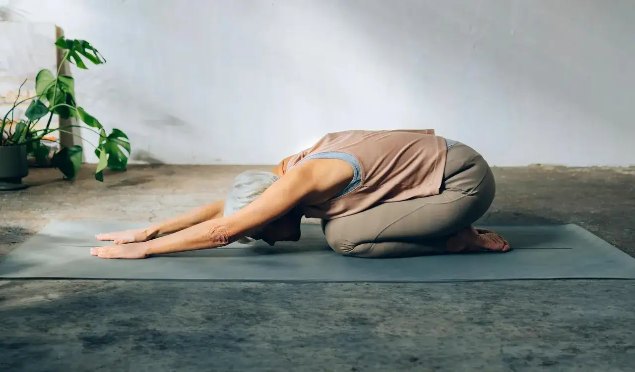 Äldre kvinna i barnets position på yogamatta. Hennes armar sträcks framåt, huvudet vilar mot golvet.