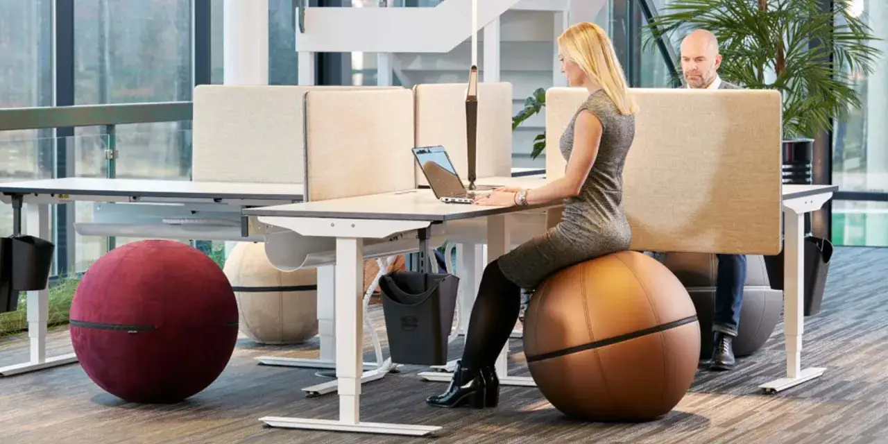 Kvinna sitter p&aring; en stor boll vid ett skrivbord i ett modernt aktivitetsbaserat kontorslandskap. En man arbetar vid ett n&auml;rliggande skrivbord.
