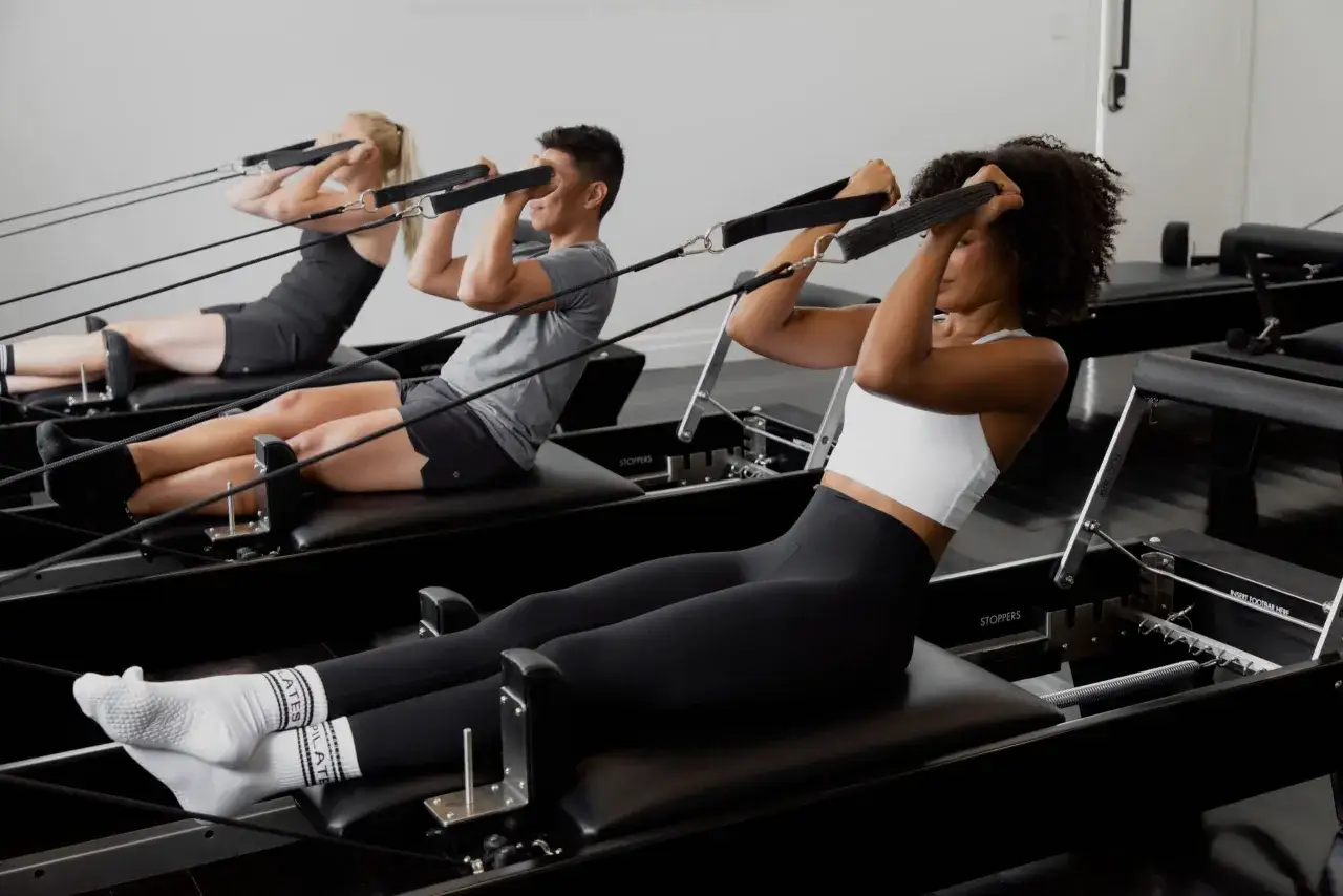 Tre personer tränar reformer pilates. De ligger på rygg och drar i handtag med rep.