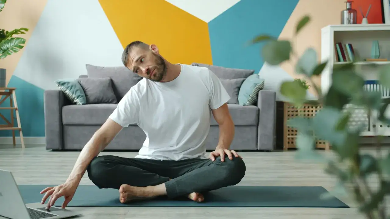 Man g&ouml;r yoga online, sitter p&aring; en matta och tittar p&aring; en laptop.