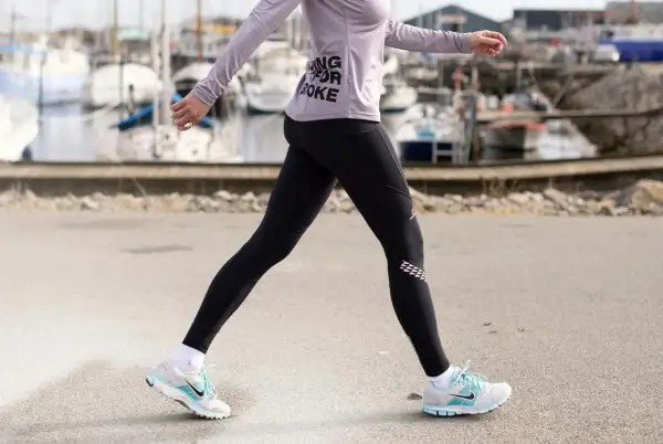 Kvinna i träningskläder går längs en kaj. Hon har på sig svarta tights och en lila tröja med texten "Running for Coke". Skorna är vita med blå detaljer. Hon tar många steg per km.
